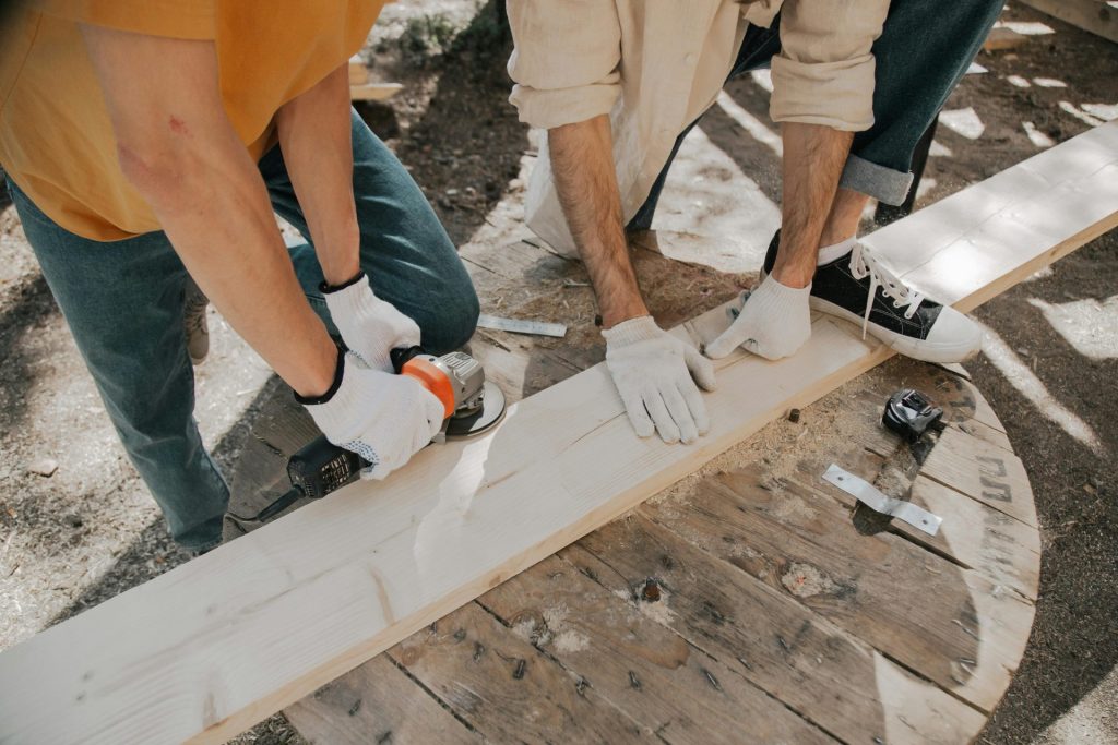 Two young carpenters using electric tools on a wooden plank outdoors. Practical DIY woodworking scene.
